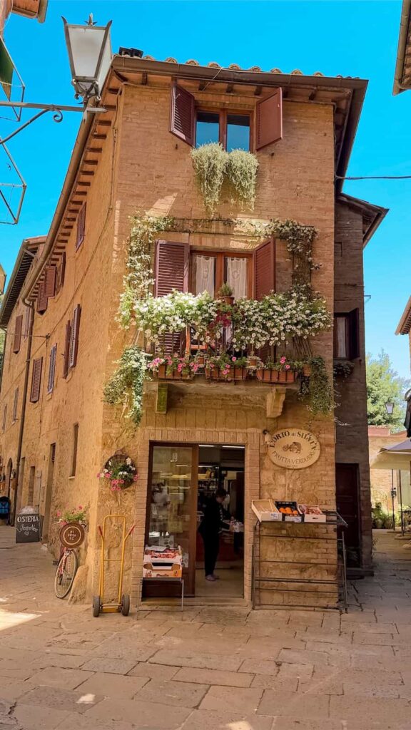A charming stone building in a Tuscan village with wooden shutters, cascading flowers from window boxes, and a small fruit and vegetable shop at street level, bathed in warm sunlight.