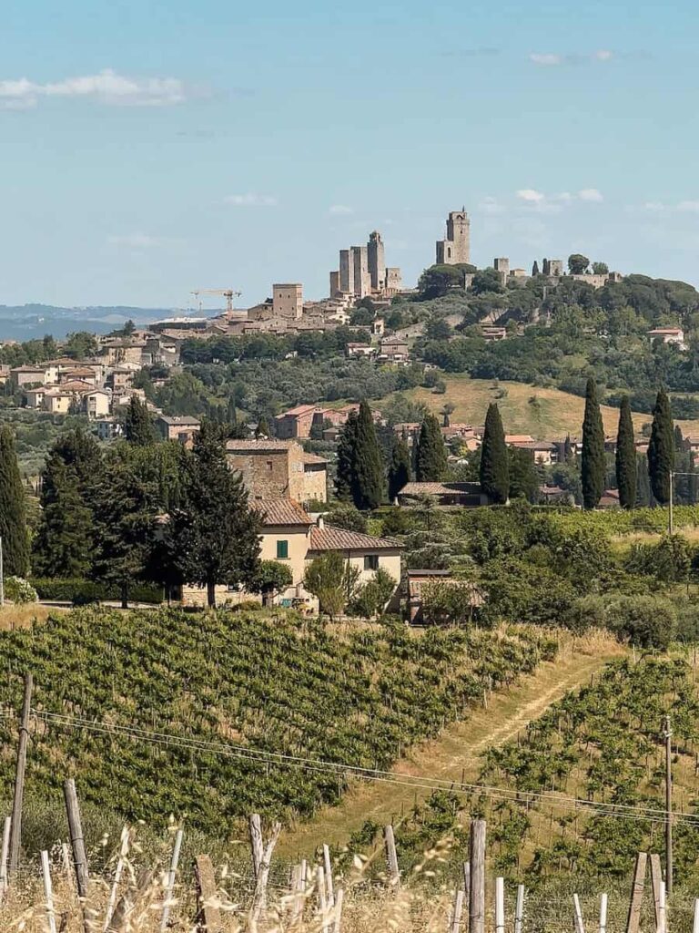 A scenic view of San Gimignano, Italy, with its medieval towers rising above a hilltop town surrounded by lush vineyards, cypress trees, and rolling Tuscan countryside under a clear blue sky.