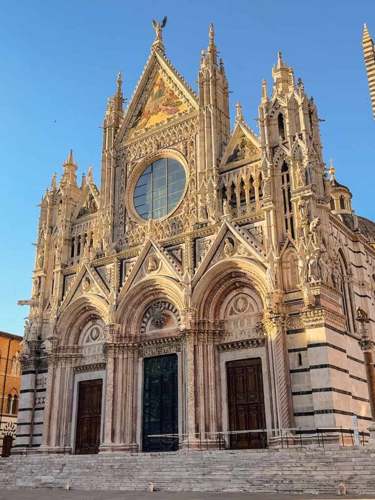 The Siena Cathedral in Italy, a stunning example of Italian Gothic architecture, with an ornate façade featuring intricate carvings, statues, striped marble patterns, and a large circular stained-glass window, illuminated by the warm light of the setting sun against a clear blue sky.