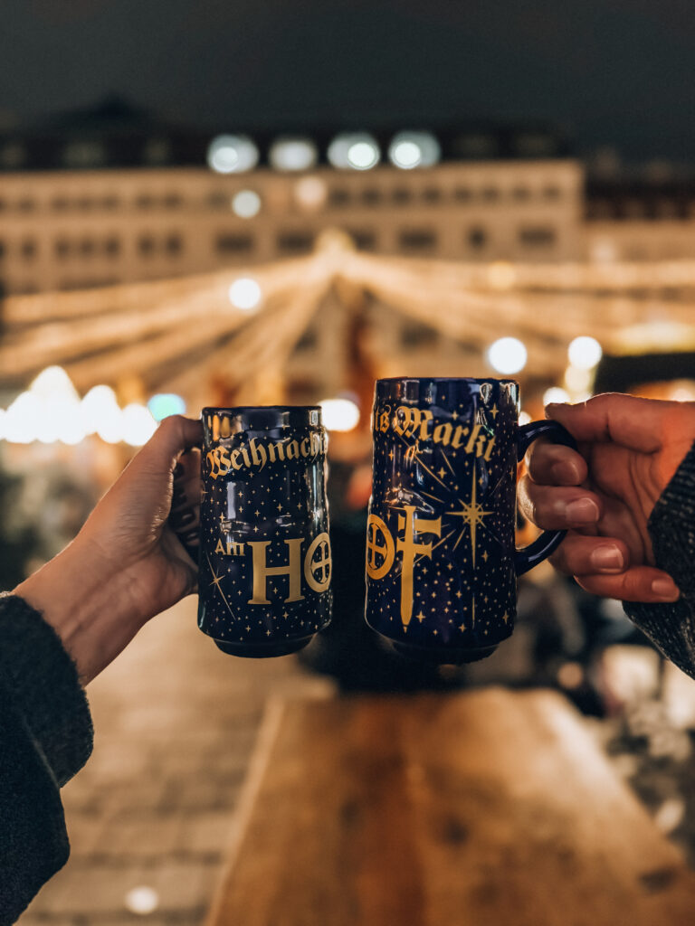 Two people clinking festive blue mugs labeled "Weihnachtsmarkt am Hof" in front of golden Christmas lights at the Am Hof Christmas Market in Vienna.

