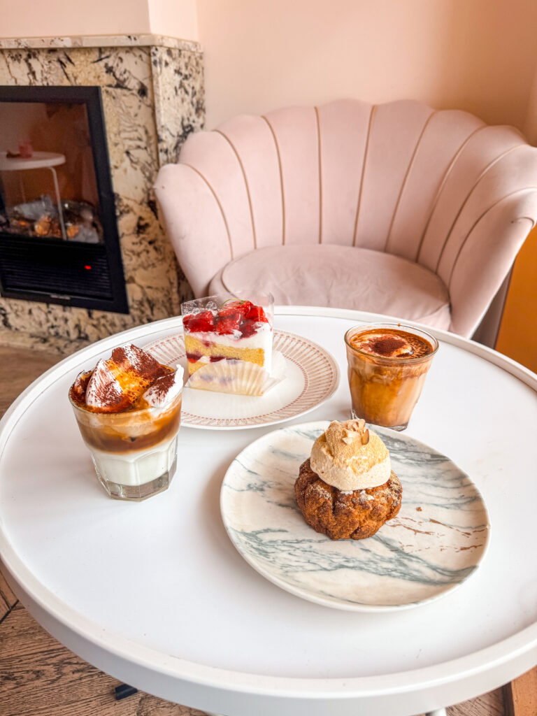 A white round table with layered cakes, whipped drinks, and a cream-topped pastry set in front of a plush pink chair and marble fireplace.