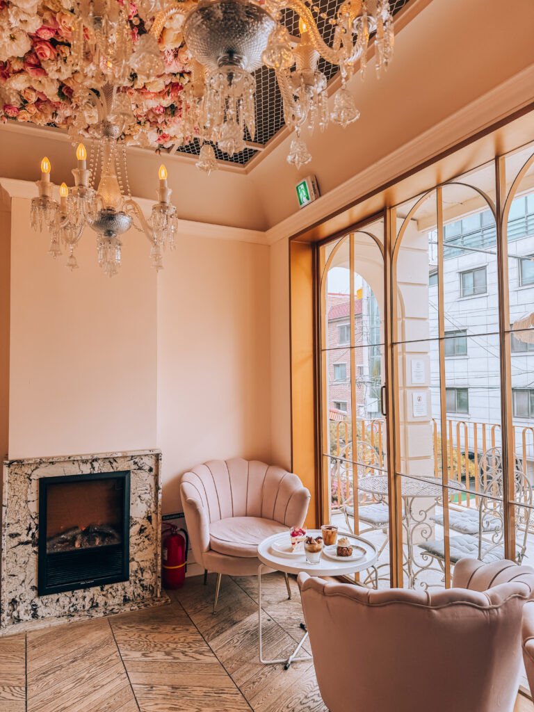 A cozy café corner with velvet chairs, a round table of desserts, and chandeliers hanging from a ceiling covered in flowers.