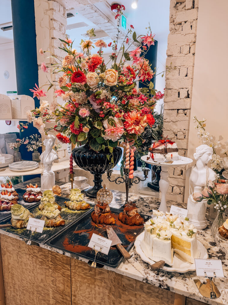 A decadent dessert table featuring matcha croissants, cakes, and pastries surrounded by floral arrangements and elegant décor in a Seoul café.