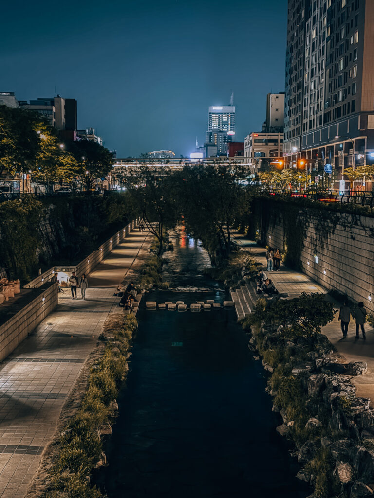 A peaceful night view of Cheonggyecheon stream, with warm lighting along the walkways and tall buildings glowing in the background suggested as one of best free things to do in Seoul Itinerary
