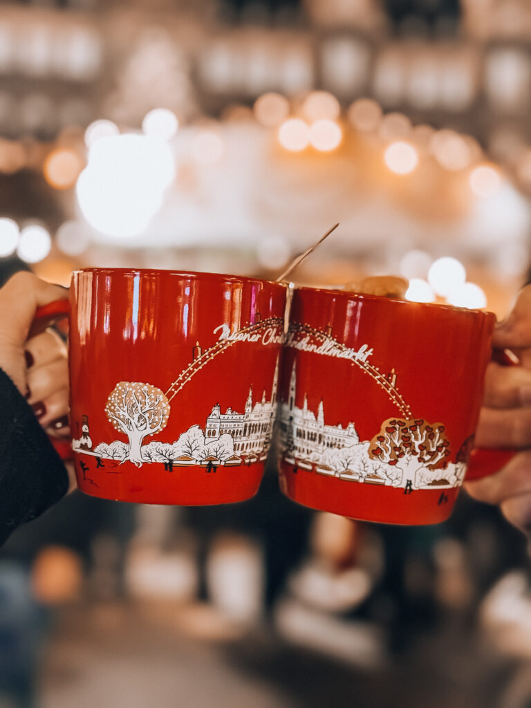 Close-up of two red souvenir mugs featuring the Vienna City Hall design, clinking together at the Rathausplatz Christmas Market with festive lights in the background.


