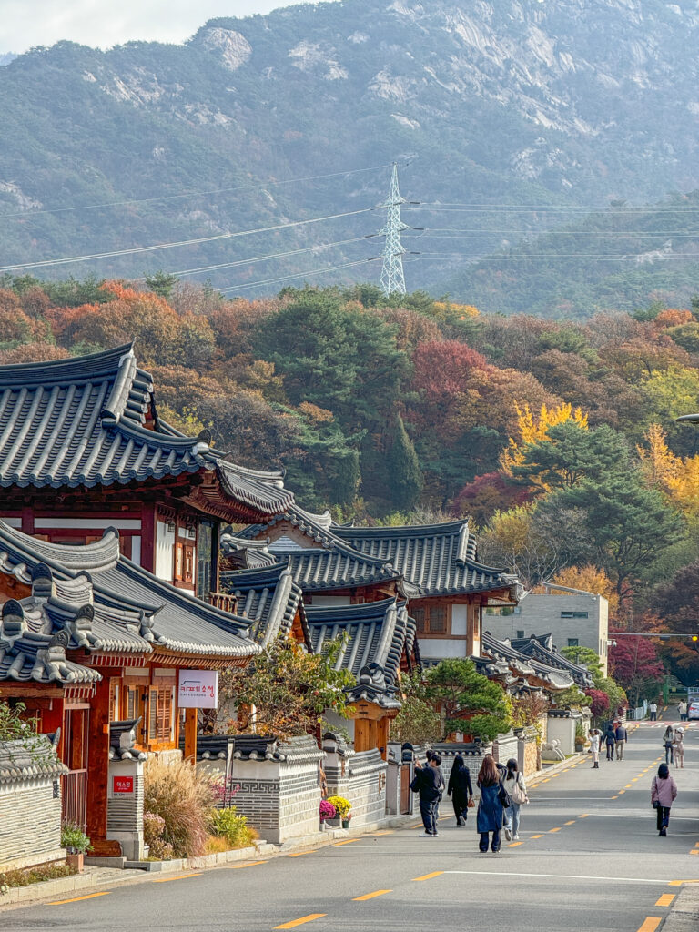 Visitors walking through Eunpyeong Hanok Village in Seoul, surrounded by traditional Korean houses with tiled roofs and vibrant autumn foliage, with mountains rising in the background.
