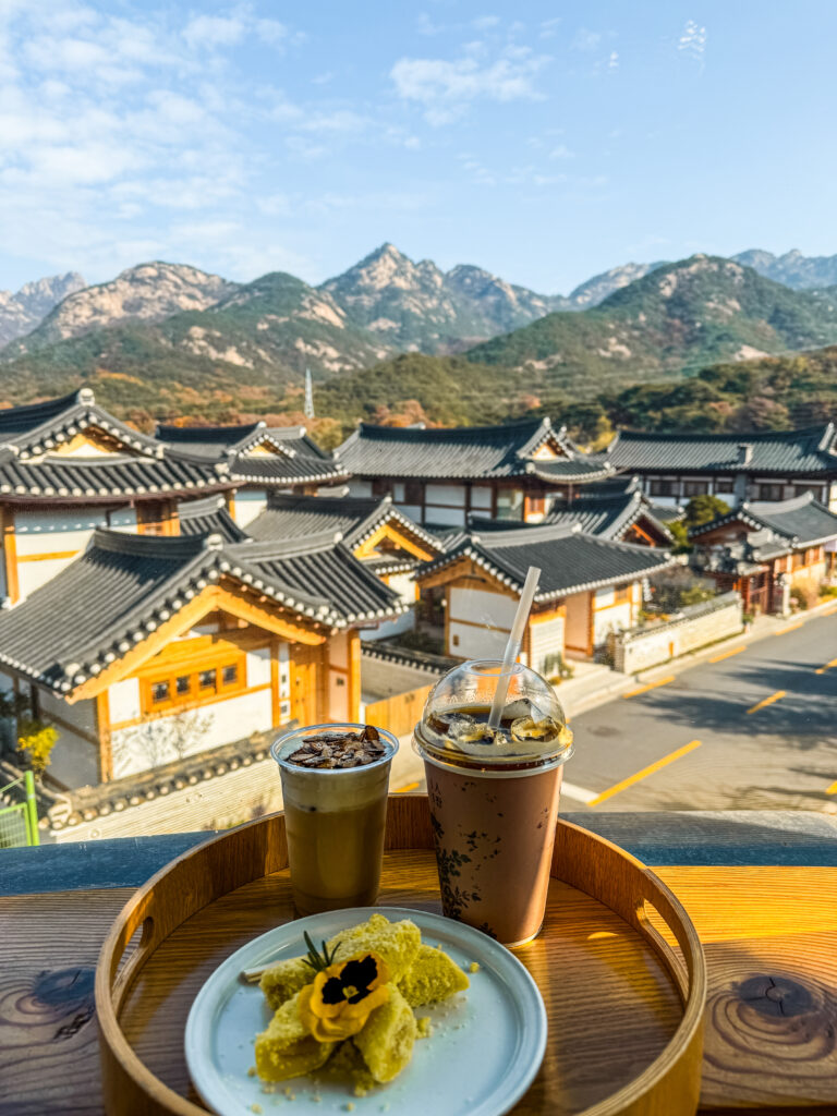 Drinks and a dessert on a wooden tray set by the window, overlooking Eunpyeong Hanok Village with traditional Korean rooftops and a stunning mountain backdrop.