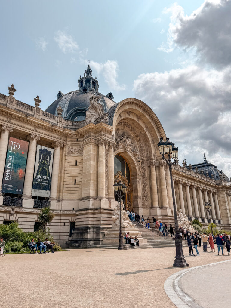 A majestic Beaux-Arts building with intricate sculptures, a domed roof, and golden doors, with visitors gathered on the steps under a partly cloudy sky