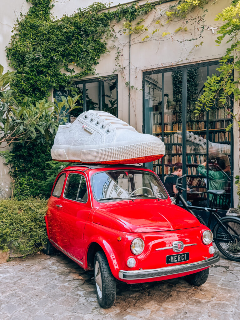 A quirky red vintage Fiat 500 with a giant white sneaker on its roof, parked in the leafy courtyard of the Merci concept store.