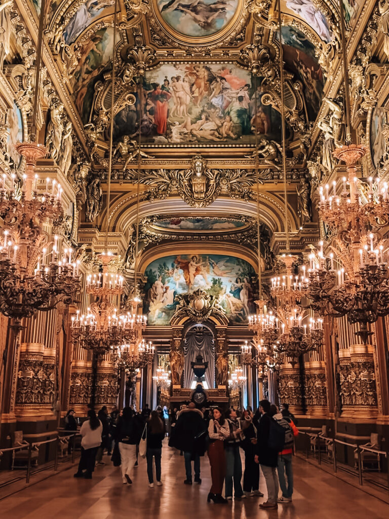 The lavish Grand Foyer of the Palais Garnier adorned with ornate chandeliers, gilded moldings, and magnificent ceiling frescoes, filled with visitors.