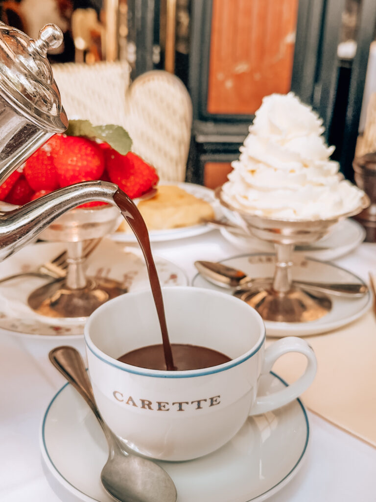 A pot of thick hot chocolate being poured into a Carette-branded cup, with strawberries, whipped cream, and elegant Parisian tableware in the background.