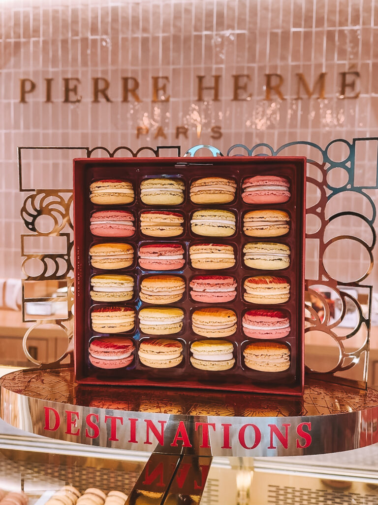 A luxury display of colorful macarons inside a Pierre Hermé boutique, perfectly arranged in a square box with a mirrored backdrop and soft lighting.