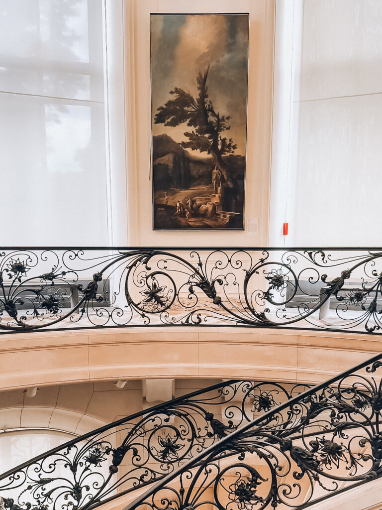 A sweeping staircase with elegant black wrought iron railings curves gracefully beneath a tall classical painting in a softly lit gallery.