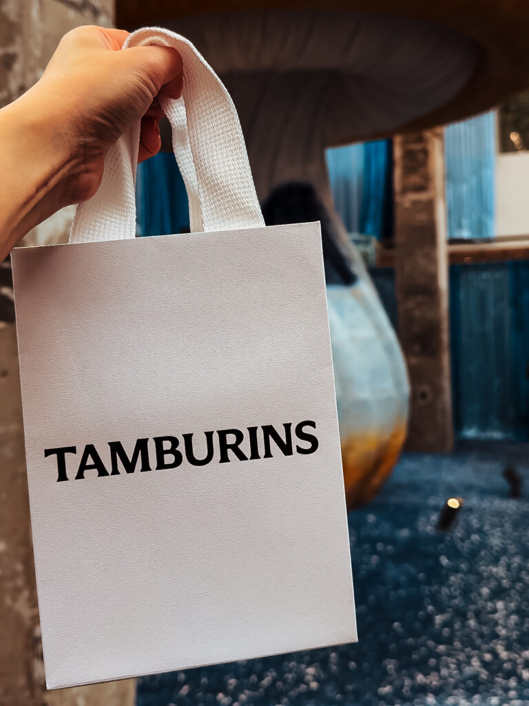 A close-up of a white Tamburins shopping bag held up against a chic, modern backdrop featuring a large mushroom-shaped art installation.