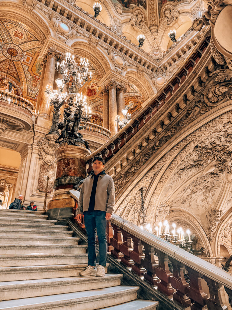 A visitor stands on the marble staircase of Palais Garnier, surrounded by elaborate stone carvings, golden details, and ornate chandeliers.