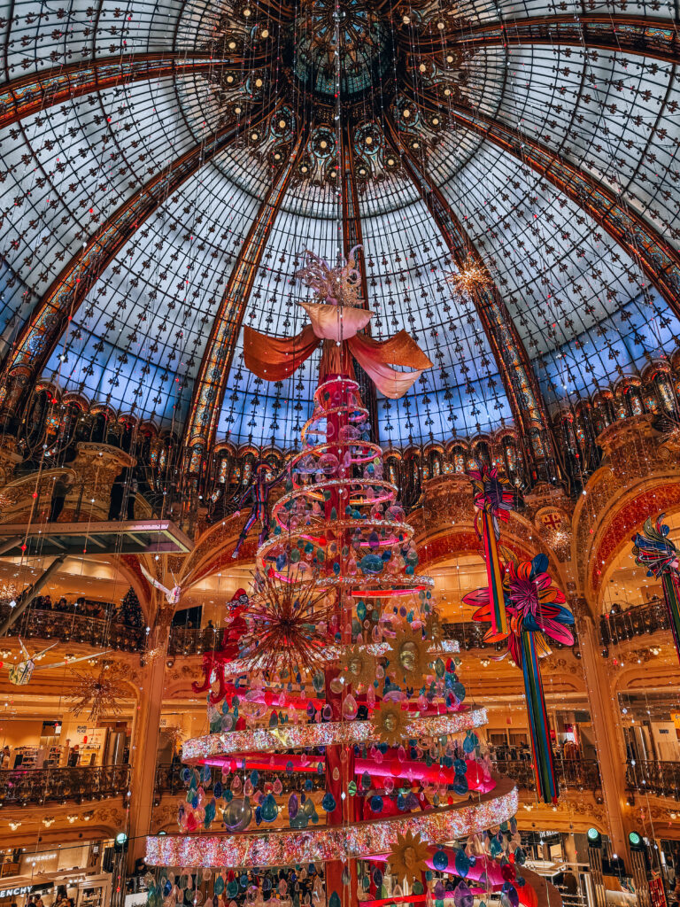 A towering, colorful Christmas tree sparkles beneath the stunning glass dome of Galeries Lafayette, with holiday ornaments suspended in the air.