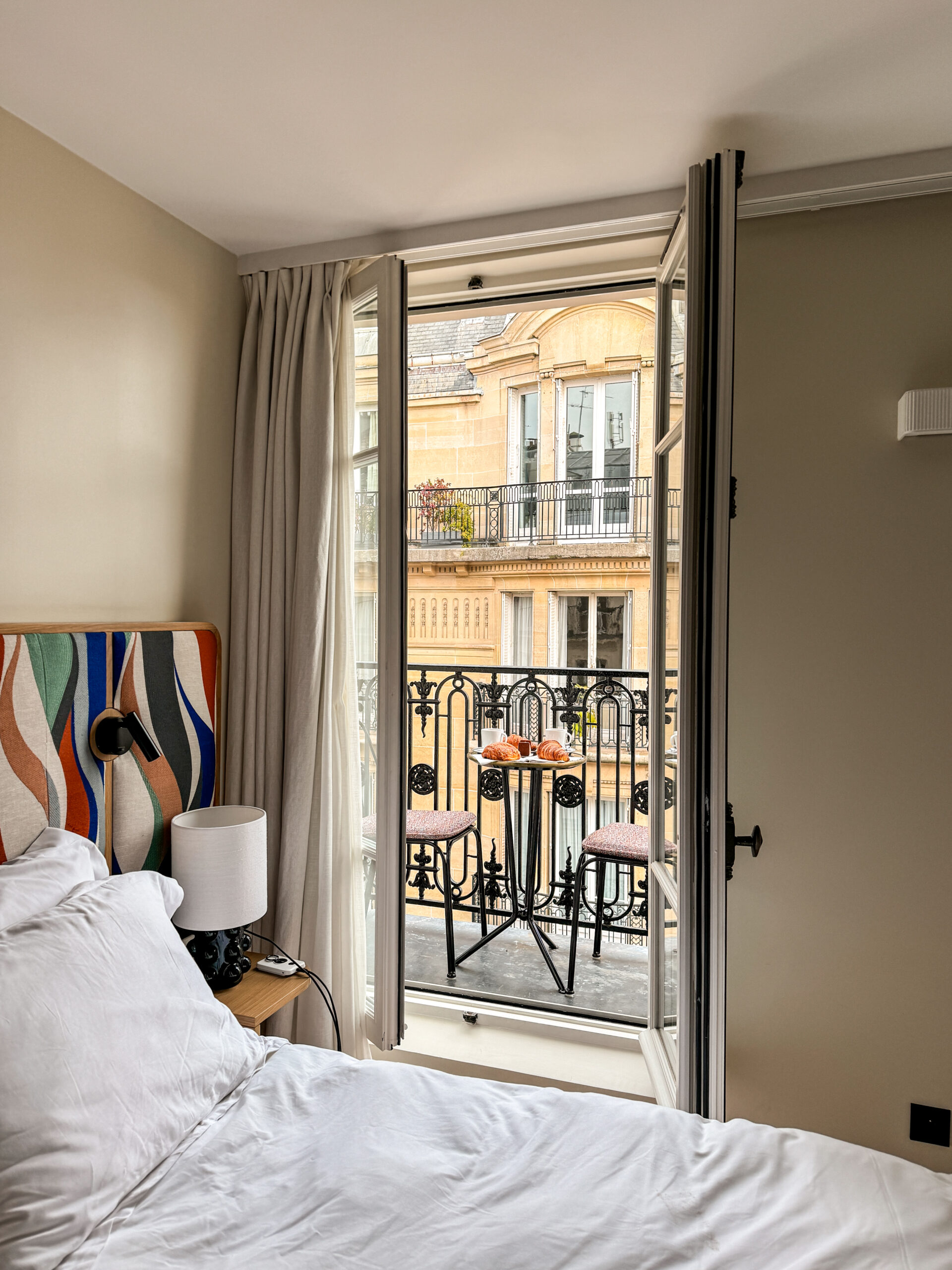 A cozy hotel room with an open balcony door revealing a small table with croissants and classic Parisian architecture in the background.