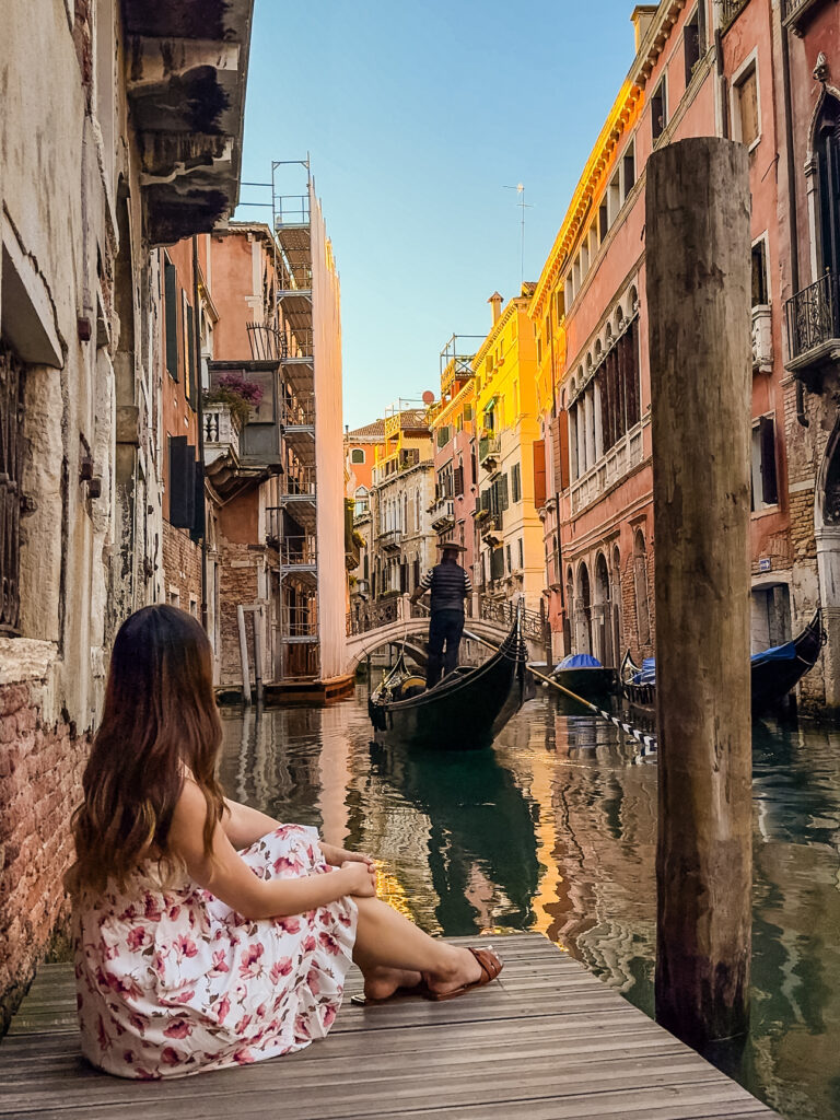 a woman sitting on the docks along canals in venice watching a gondola
