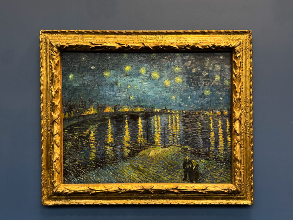 A framed oil painting of Van Gogh’s “Starry Night Over the Rhône” displayed on a navy blue wall, glowing with rich yellows and deep blues.