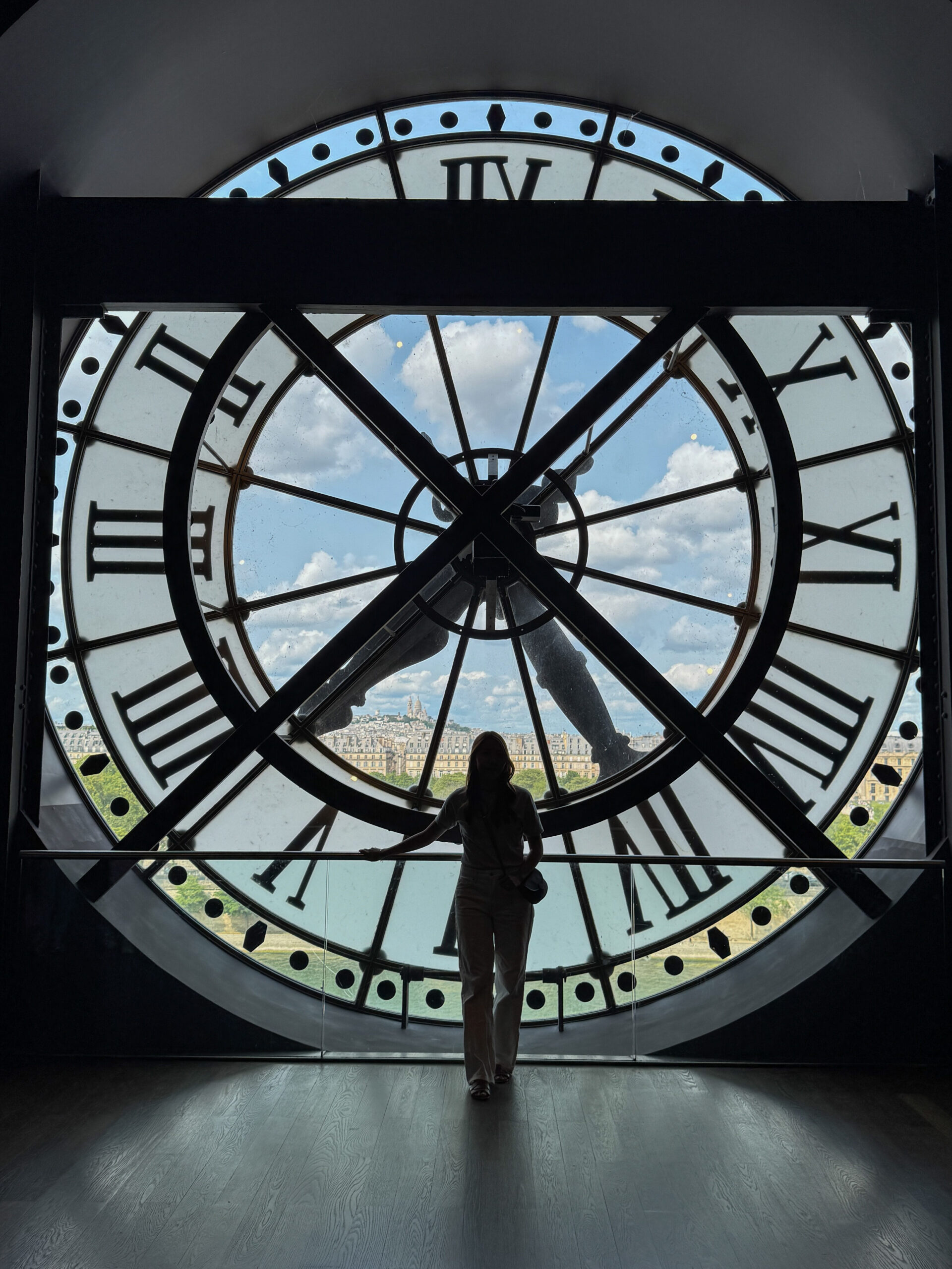 A silhouette of a woman standing inside the giant clock window of Musée d'Orsay, with a panoramic view of Paris visible through the glass.