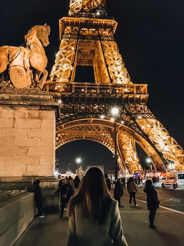 The Eiffel Tower illuminated against the night sky as crowds stroll beneath its sparkling lights, with a stone horse sculpture in the foreground.