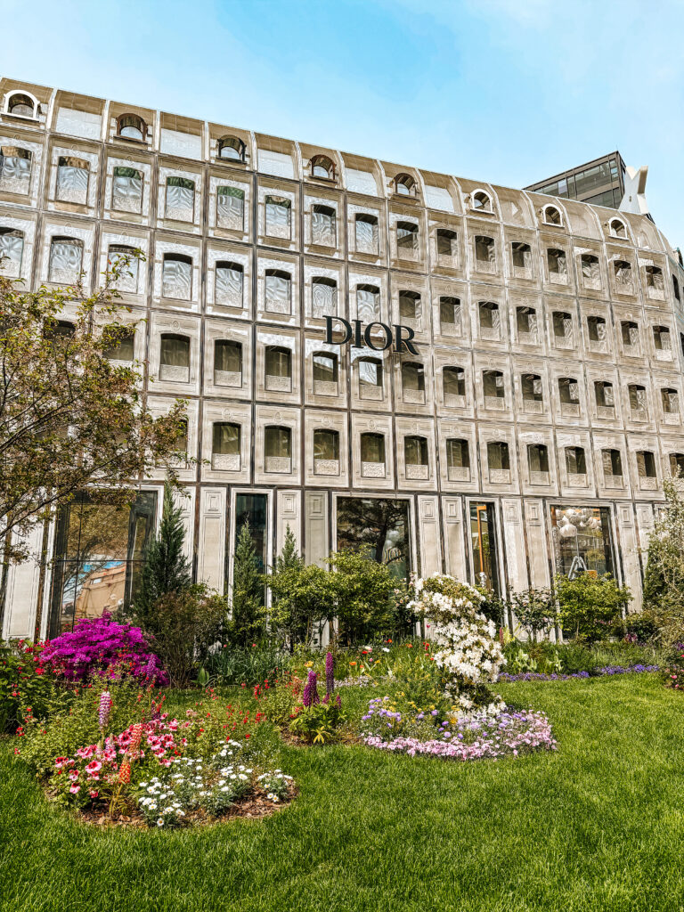 The elegant exterior of the Dior flagship store in Seoul, framed by a lush, manicured garden filled with colorful spring flowers.