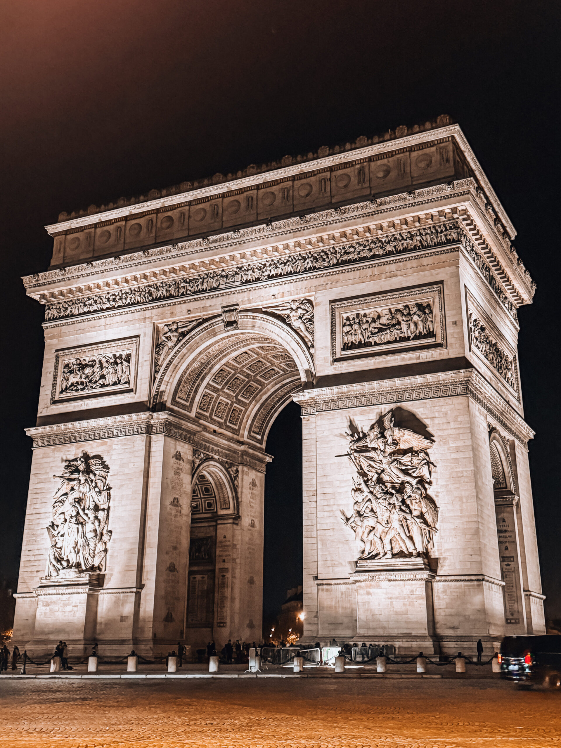 The Arc de Triomphe lit dramatically under a dark sky, with intricate carvings and people gathered at its base.