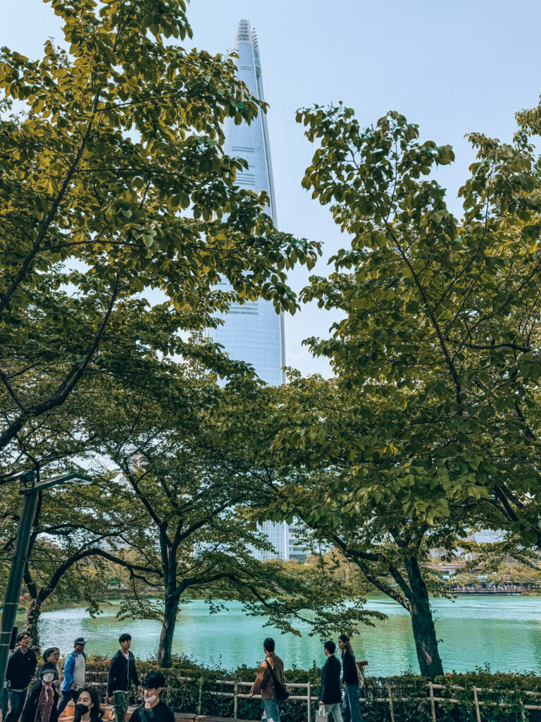 Lotte World Tower peeking through lush green trees by the turquoise waters of Seokchon Lake, with people strolling along the lakeside path.