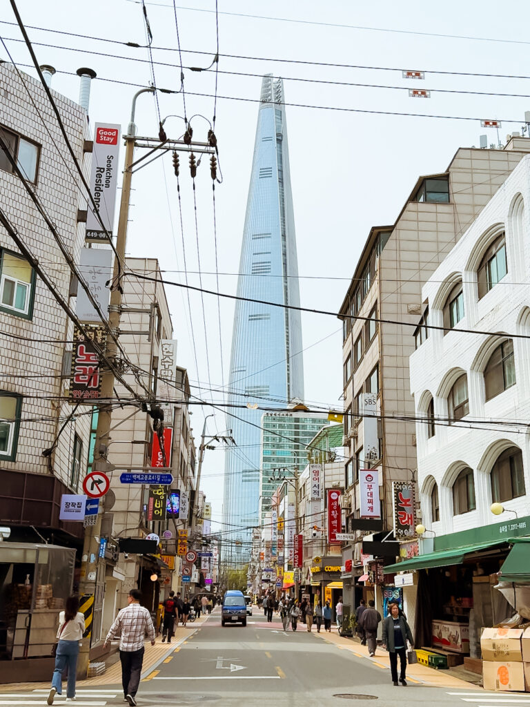 A bustling Seoul street scene framed by low-rise buildings, with the towering Lotte World Tower rising in the distance under a pale sky.