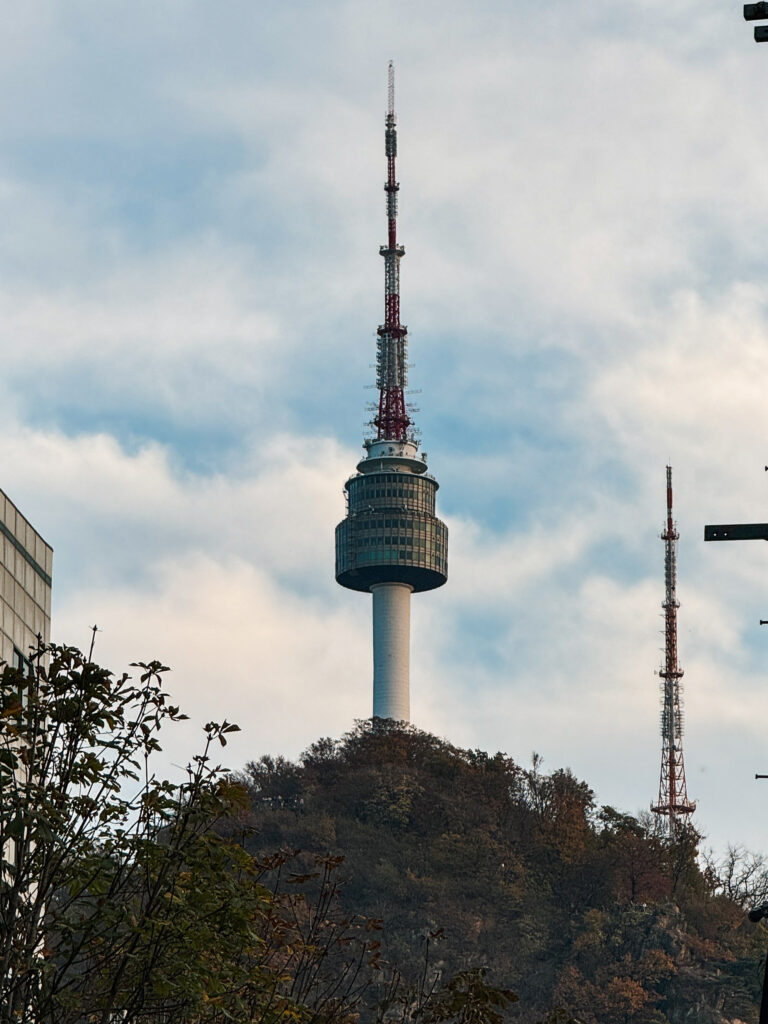 N Seoul Tower standing tall atop Namsan Mountain, framed by autumn foliage and a dramatic cloudy sky.