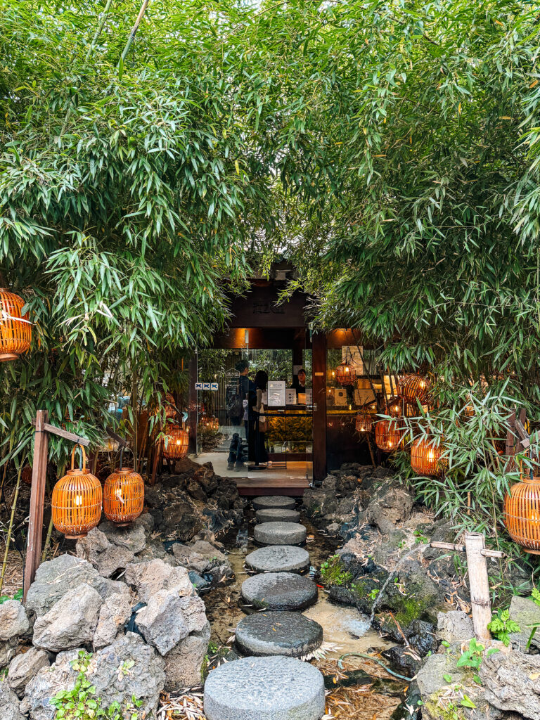A serene entrance path of round stone steps over a small stream, surrounded by lush bamboo and warm lantern lighting at Cheongsudang café.
