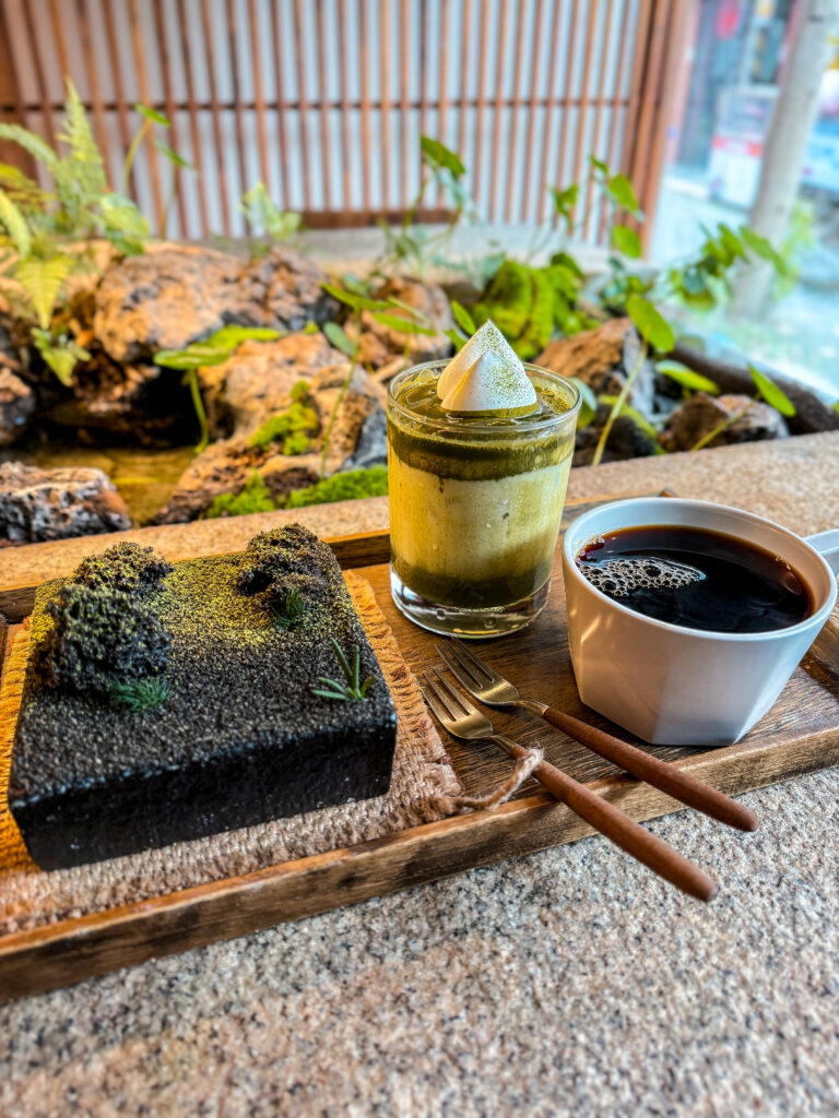 A beautifully presented dessert and drink set at a Korean café, featuring a matcha dessert styled like a zen garden, an iced matcha latte with whipped topping, and a black coffee in a white ceramic cup, placed on a wooden tray with a stone and moss garden in the background.