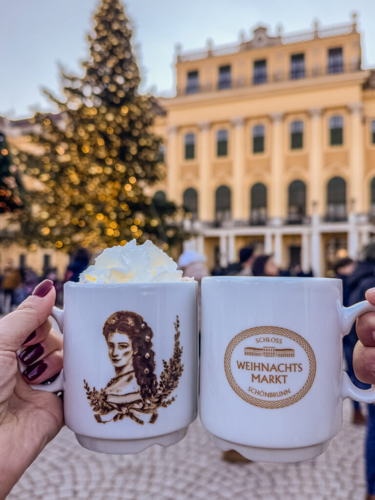 Close-up of two white mugs from the Schönbrunn Palace Christmas Marke. One featuring Empress Sisi and the other with the market’s logo, held up in front of the decorated palace and Christmas tree.
