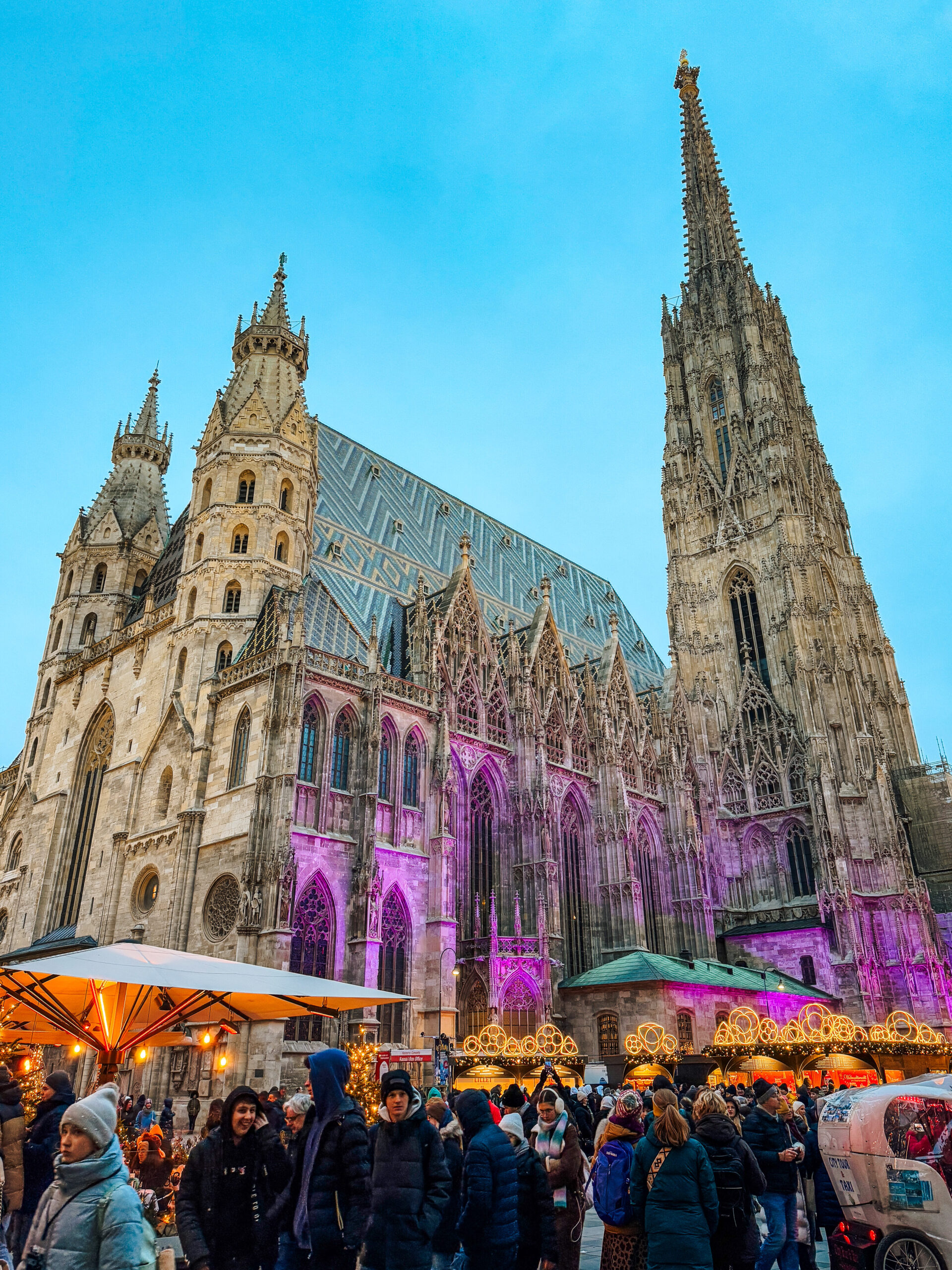 Crowds walking through the Stephansplatz Christmas Market in Vienna at dusk, with St. Stephen’s Cathedral illuminated in purple lights behind festive stalls and holiday decorations.