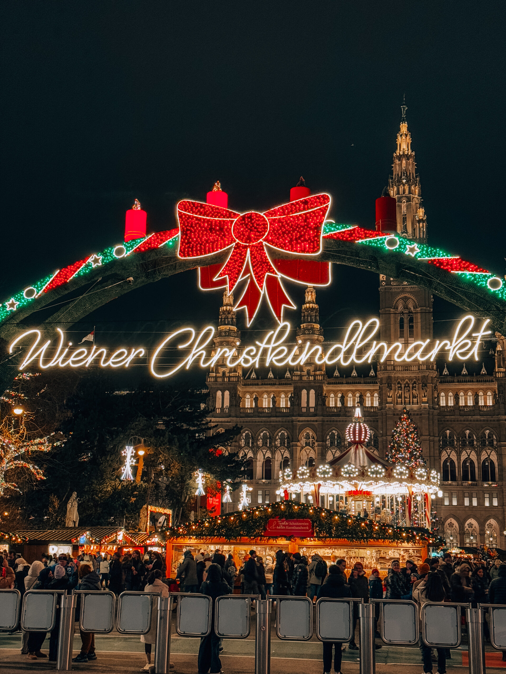 Nighttime scene of the Wiener Christkindlmarkt at Rathausplatz in Vienna, with a glowing red bow and candle arch, illuminated City Hall, and carousel surrounded by Christmas lights and market stalls. This is the most famous of the Christmas markets in Vienna. 