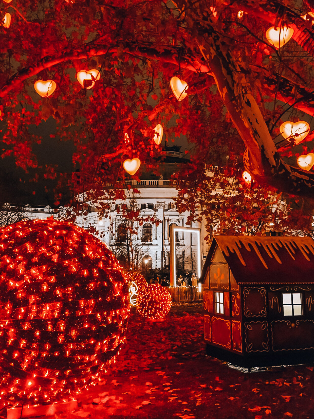 Enchanting night scene from Vienna’s Rathauspark with glowing red heart lights hanging from trees, oversized Christmas baubles, and a gingerbread-style hut illuminated in festive red.