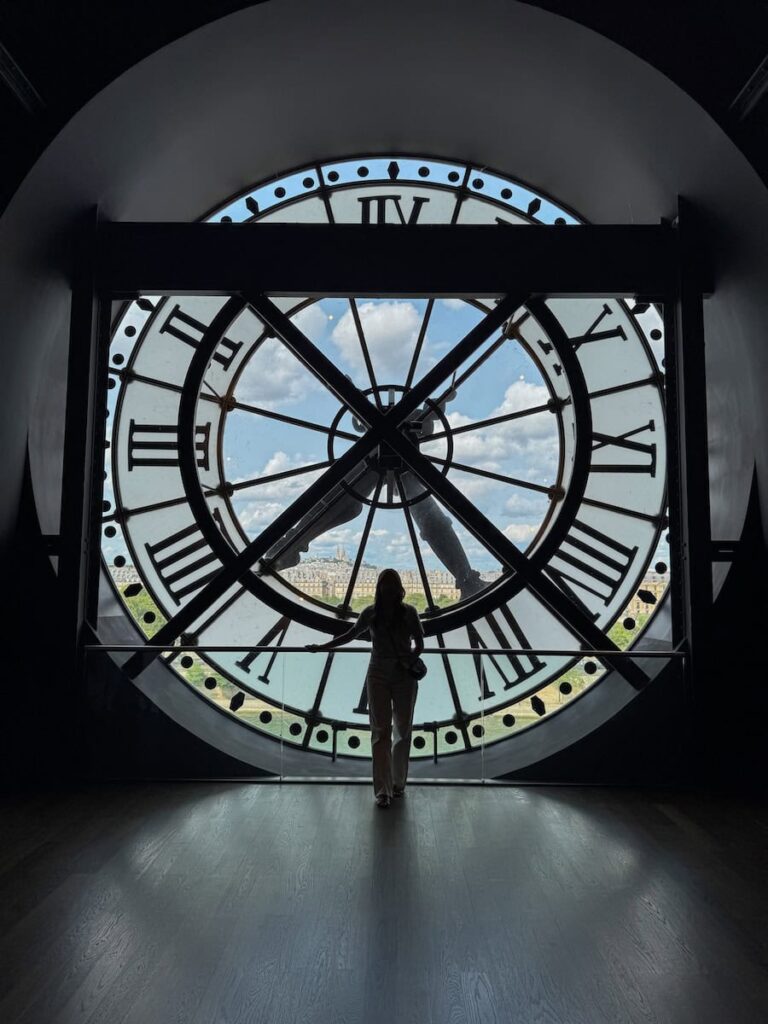 A silhouette of a woman standing inside the giant clock window of Musée d'Orsay, with a panoramic view of Paris visible through the glass.