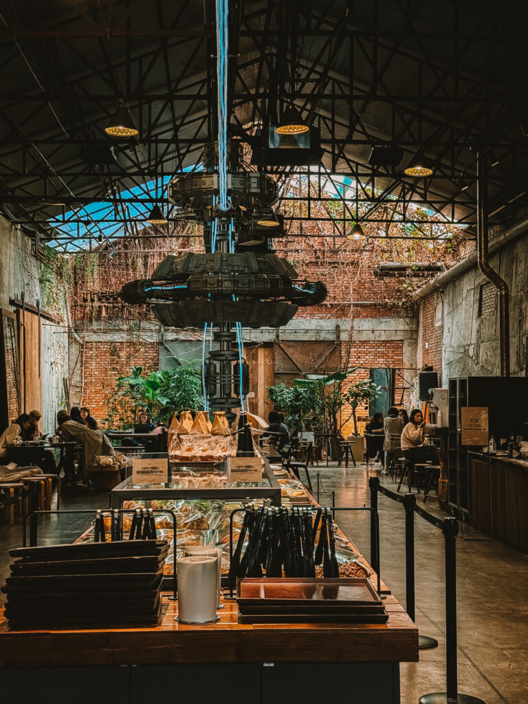 Interior of a spacious industrial-style Daelim Changgo Cafe in Seoul, featuring exposed brick walls, high ceilings with steel beams, lush indoor plants, and a futuristic central art installation above a self-serve pastry counter.