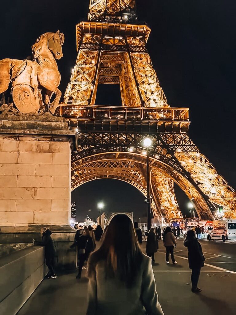 The Eiffel Tower illuminated against the night sky as crowds stroll beneath its sparkling lights, with a stone horse sculpture in the foreground.