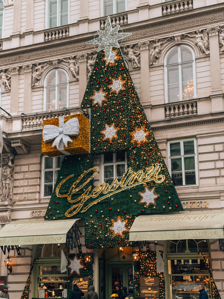 Festive Christmas tree-shaped decoration with sparkling stars and a gift box adorning the historic Gerstner Café-Restaurant façade in Vienna during the holiday season.

