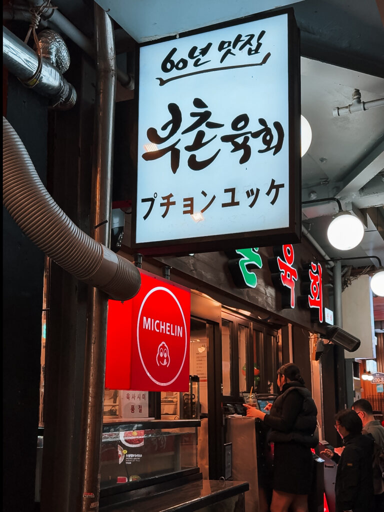 A Michelin-recognized Korean street food stall called Buchon Yukhoe with a bright white and red sign in Hangul and Japanese, located in a bustling food alley in Seoul.
