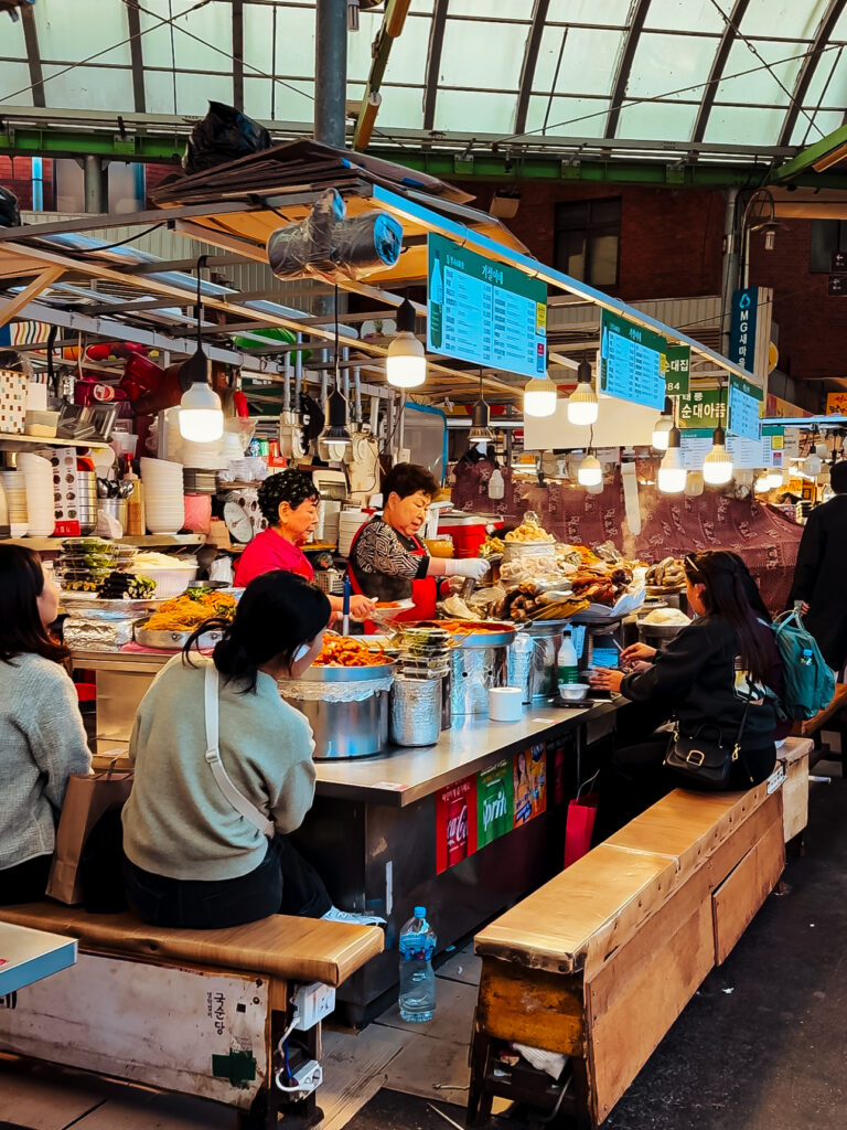 Locals and tourists seated at a vibrant food stall in Gwangjang Market, enjoying Korean street food served by vendors in a lively market atmosphere
