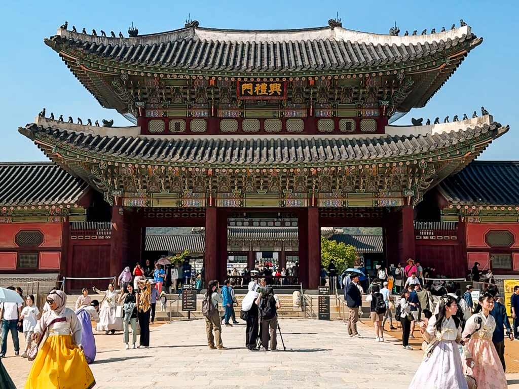The grand main gate of Gyeongbokgung Palace bustling with visitors, many dressed in hanbok, beneath the intricately detailed roof structure.