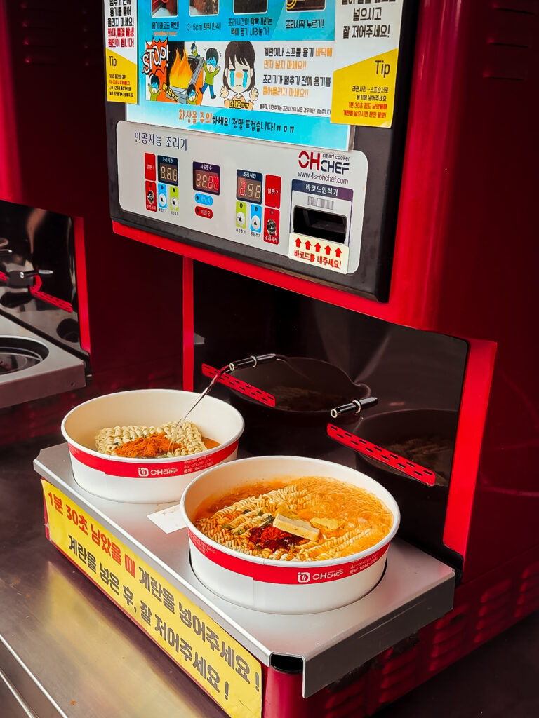 Close-up of an automated ramen cooking machine in Seoul preparing instant noodles in paper bowls, a popular and quirky local food experience.
