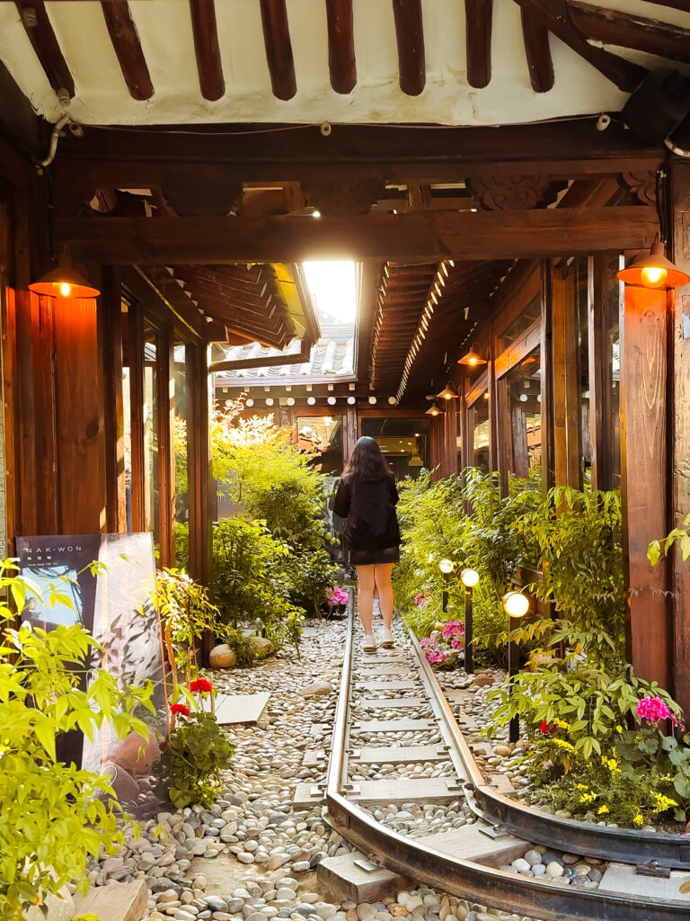 nakwon station cafe in ikseondong, Atmospheric view of a hanok-style café in Seoul with a miniature railway track pathway surrounded by plants and glowing lights.
