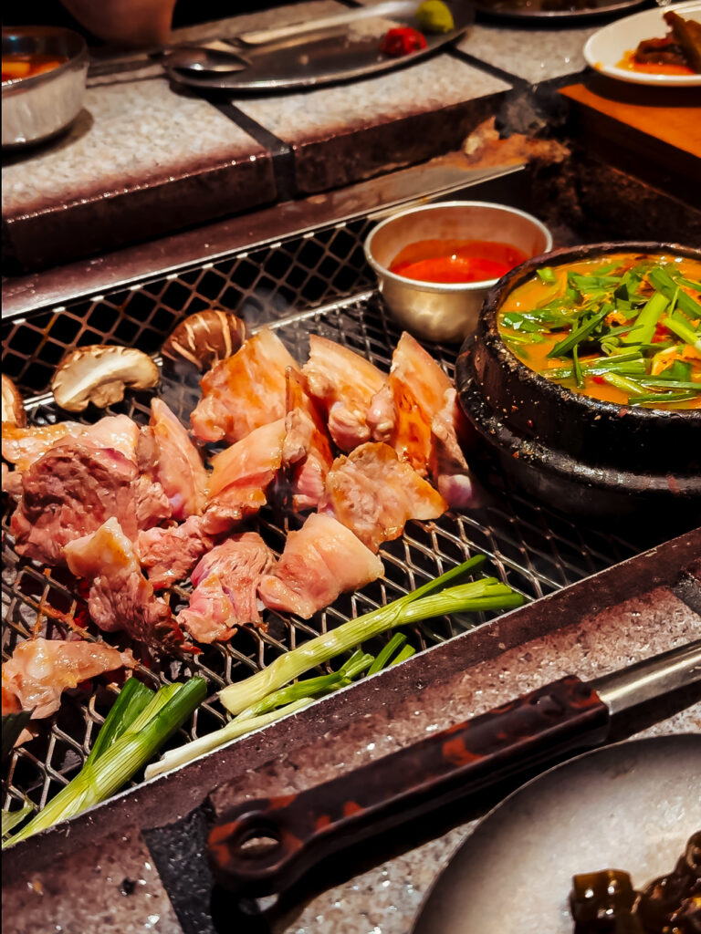 Close-up of sizzling pieces of pork belly and green onions grilling over charcoal, accompanied by a bubbling stone pot of Korean stew. 

