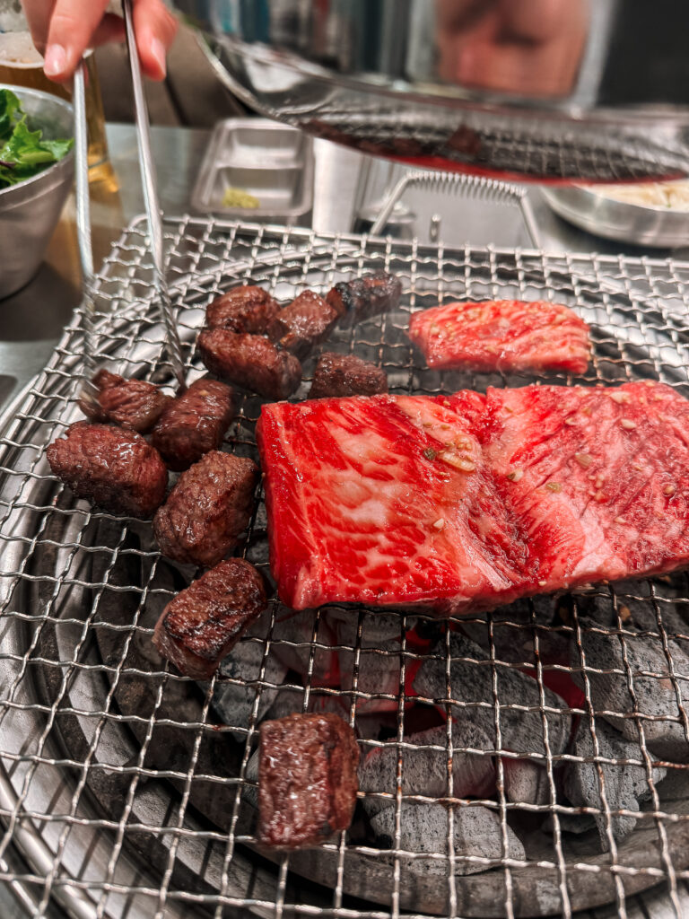 Thick slices of marbled beef and juicy meat cubes grilling on a wire mesh over glowing coals at a Korean BBQ restaurant.