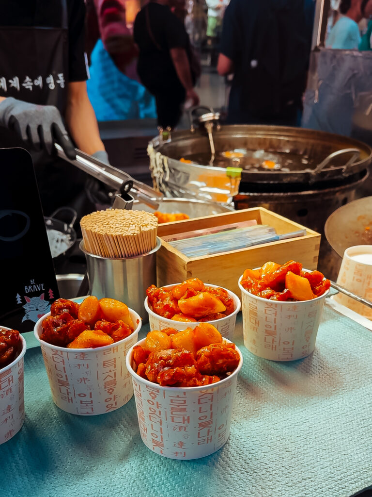 Paper cups filled with spicy Korean tteokbokki and fried snacks displayed on a street food stall in Seoul.