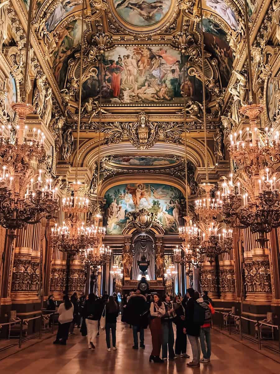The lavish Grand Foyer of the Palais Garnier adorned with ornate chandeliers, gilded moldings, and magnificent ceiling frescoes, filled with visitors.
