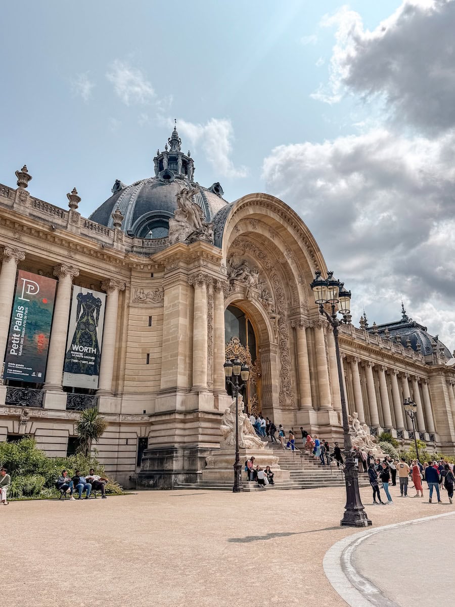 A majestic Beaux-Arts building with intricate sculptures, a domed roof, and golden doors, with visitors gathered on the steps under a partly cloudy sky
